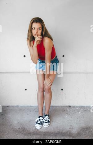 Teen girl in red top, denim mini skirt and sneakers doing silence sign with finger and eyes wide opened against a concrete wall Stock Photo
