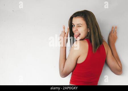 Funny teen girl in short red top making victory sign with fingers, winking and stiking out her tongue against a concrete wall Stock Photo