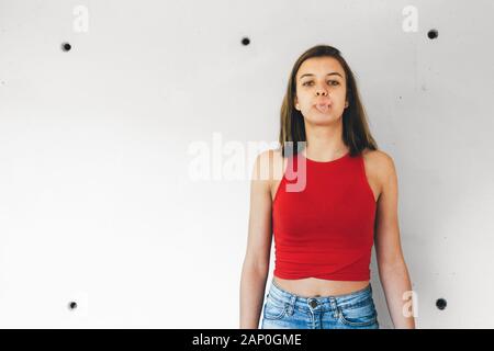 Rebel teen girl wearing a short red top and a denim mini skirt triying to blowing a chewing gum bubble against a concrete wall Stock Photo