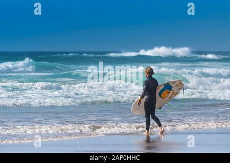 A male surfer carrying his surfboard and looking out to sea at Fistral in Newquay in Cornwall. Stock Photo