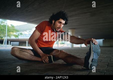 Young man athlete with earphone in his ears warming up and stretching his legs before running in the city street under the bridge Stock Photo
