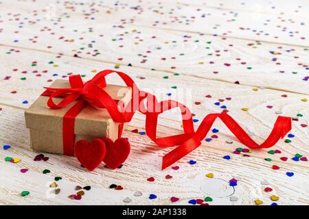 Brown gift box with red ribbon bow and two felt valentine hearts on a white wood surface with heart shaped foil confetti. Valentine greeting card. Stock Photo
