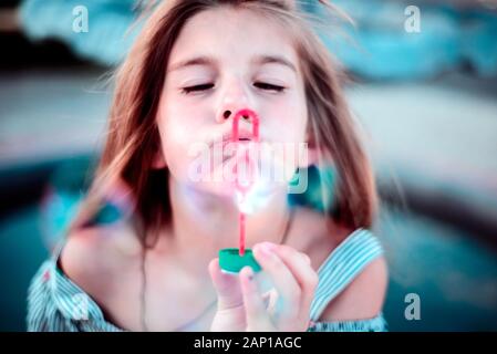 Portrait of a beautiful little girl blowing soap bubbles. A child plays with bubbles, on a green background. Outdoor Stock Photo