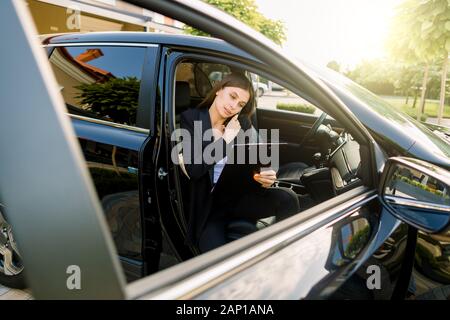 Side angle view of young pretty businesswoman, sitting inside her car on passenger seat, while using a digital tablet and talking mobile phone Stock Photo