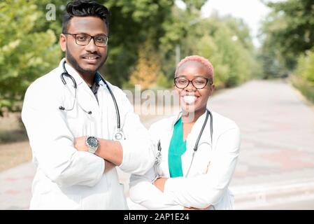 portrait of young doctors. Indian guy, black girl. People in white coats, glasses, with stethoscopes. Outdoor in Stock Photo
