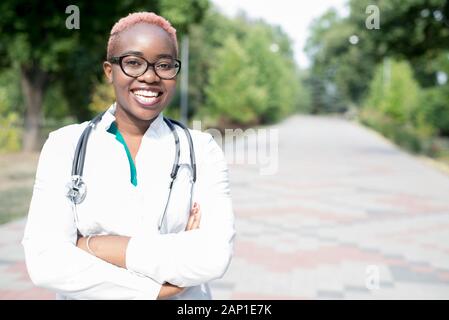 portrait of a young black girl, a doctor. Woman in a white coat, smiling, wearing glasses, with a stethoscope. Outdoor in Stock Photo