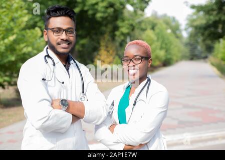 portrait of young doctors. Indian guy, black girl. People in white coats, glasses, with stethoscopes. Outdoor in Stock Photo