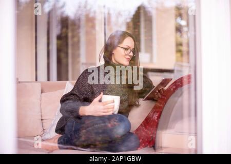 Shot of pretty young woman sitting behind glass window in the living room while using digital tablet and drinking tea. Stock Photo