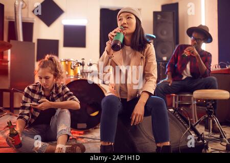 Multi-ethnic group of young people in music studio focus on Asian young woman drinking beet in foreground Stock Photo