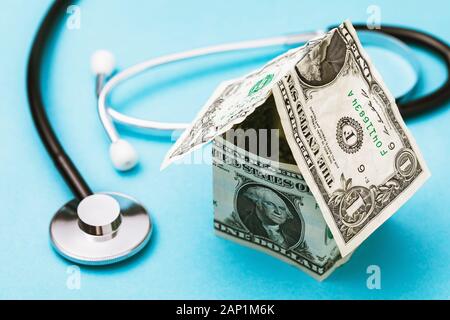 House made of dollars and a medical stethoscope on a blue background. Concept on the cost of a call to a doctor at home Stock Photo