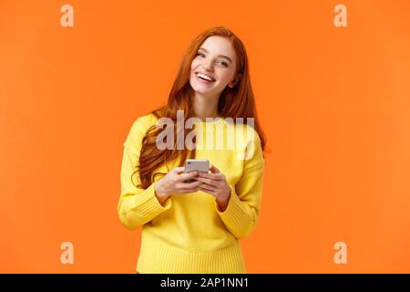 Girl searching gifts for holidays online. Carefree happy redhead female student using smartphone app, laughing joyfully, holding mobile phone Stock Photo