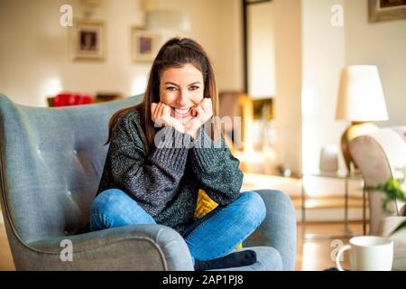 Shot of happy woman with hands on chin sitting in the armchair and relaxing at home. Stock Photo