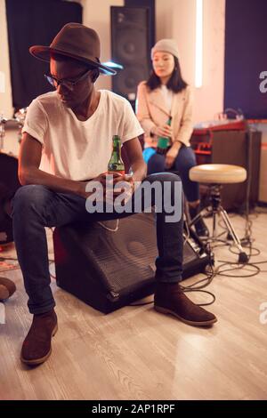 Full length portrait of trendy African man wearing hat and holding beer bottle while relaxing in music studio Stock Photo