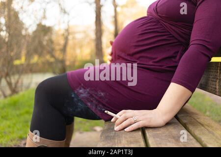 Close-up on pregnant woman with smoking cigarette in her hand, sitting on the bench in park. Bad habits during pregnancy and the risk for child health Stock Photo