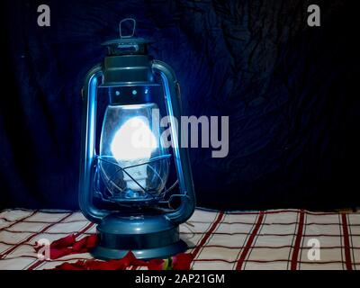 a hurricane lamp on a patterned cloth with red rose petals spread at its base setting a romantic scene. Stock Photo