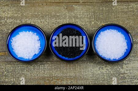 Close up of three isolated blue bowls with different kind of salt on wood table (white flakes and grains, black Hawaii salt) Stock Photo