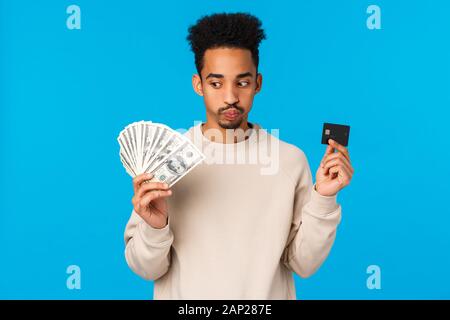 Indecisive and troubled young african-american guy facing tough decision, pouting, exhale hard making choice, holding money and credit card, consider Stock Photo
