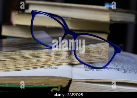 View on isolated stack of old yellowed books on wood table with blue reading glasses and open book Stock Photo