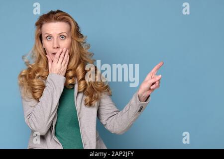 Amazed excited mature woman pointing at copy space aside, happy surprised middle aged lady looking at camera on blue wall. Studio shot Stock Photo