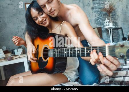 Mixed Race Couple. Young man teaching woman playing guitar sitting on bed together cllose-up laughing playful Stock Photo