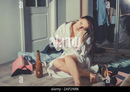 portrait of a beautiful young girl in a home white coat. He sits on the floor in a room with scattered things and pours wine into a glass. For a long Stock Photo