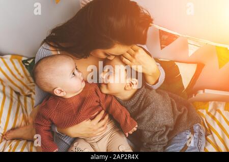 Young beautiful mother with her children on the bed, tightly hugs her son and little daughter, kisses her forehead, children laugh Stock Photo