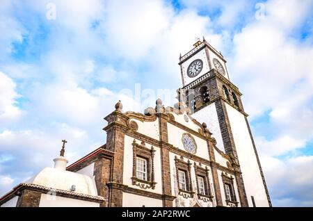 The outdoor facade of St. Sebastian Church, Igreja Matriz de Sao Sebastiao, in Ponta Delgada, Azores, Portugal. White clock tower from below with blue sky and clouds above. Sunset light, golden hour. Stock Photo