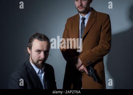 Portrait of two men in business suits, one of them is sitting, the other is holding a gun. On a gray background. Dramatic light. Artistic setting. The Stock Photo