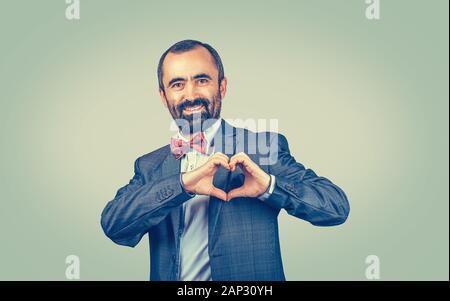 Handsome smiling mature middle-aged man makes heart shape with fingers, hands, isolated on green yellow wall background. Positive human emotion facial Stock Photo