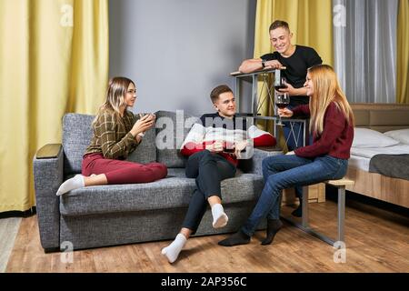 A group of young men and women drink alcohol at a college dorm party. Drunk on the university campus, undergraduates are sitting in the bedroom with g Stock Photo