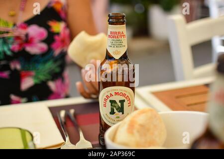 A woman in a flower print top eats a bread roll and enjoys a glass bottle of beer at an outdoor patio in Italy. Stock Photo