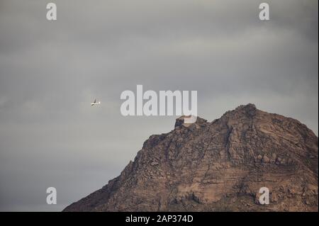From below wonderful view of white airplane flying in sky under lush gray clouds above brown rocky mountain summit Stock Photo
