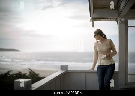 Young woman standing with a tea cup on a balcony by the sea. Stock Photo