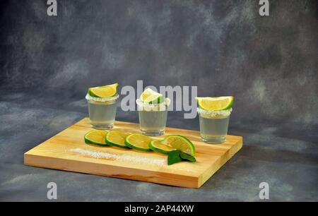 Three shots of tequila with salt and lime slices on a wooden stand next to slices of citrus and a pinch of salt. Close-up. Stock Photo