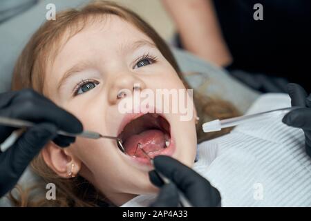 Portrait of pretty bold female child sitting in dental chair with open mouth while professional doctor in rubber gloves carefully examining condition of teeth. Concept of dentistry Stock Photo