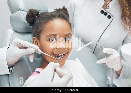 Happy afro american girl sitting in stomatologist chair with open mouth while professional dentist doing regular check up of teeth using dental probe and mirror. Female nurse assisting. Stock Photo