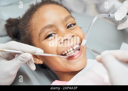 Charming little afro american girl sitting in dental chair, smiling and looking at camera during medical treatment at modern clinic. Concept of health care ad pediatrics Stock Photo