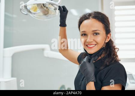 Portrait of beautiful female dentist in black medical uniform smiling and looking at camera with blur background of dental cabinet. Happy confident woman at working place. Stock Photo