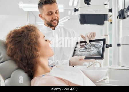 Side view of beautiful girl with curly hair sitting in dental chair and looking at electronic device that holding handsome male doctor. Professional dentist showing x-ray pictures of teeth to client Stock Photo