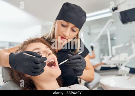 Female dentist in medical uniform, black hat, glasses and rubber gloves doing regular oral check up with mouth mirror and probe. Beautiful female patient with curly hair in dental clinic. Stock Photo