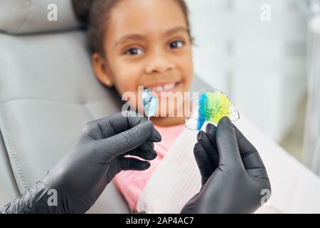 Close up of female hands in black rubber gloves holding colorful braces for alignment of teeth. Dentist showing metal teeth retainers to cute african girl sitting in dental chair Stock Photo