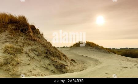 Two large sandy dunes, the sun on pastel colored sky and the roof of a house in the distance. Stock Photo