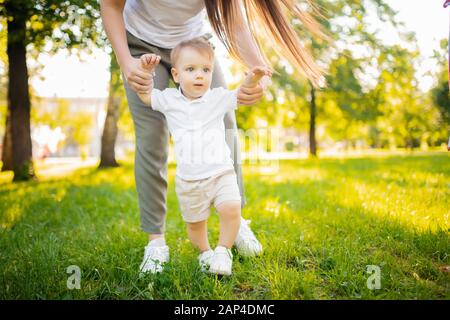 Child baby takes first step with help of mom in park. Concept parents teach walking Stock Photo