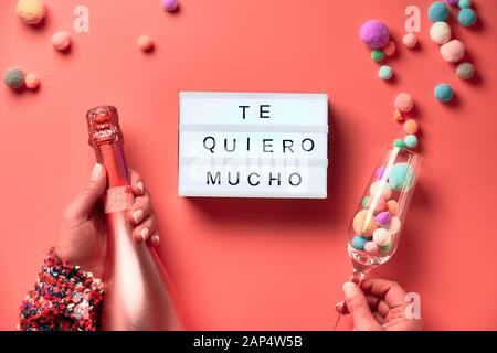 Text 'Te quero mucho' means I love you so much in English. Lightboard with text in Latino female hands, flat lay on pink background. Bottle of champag Stock Photo