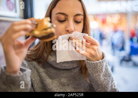Beautiful young cute girl in jumper cleans her mouth after a big bite and enjoys fast food hamburger while sittting in a cafe. Stock Photo
