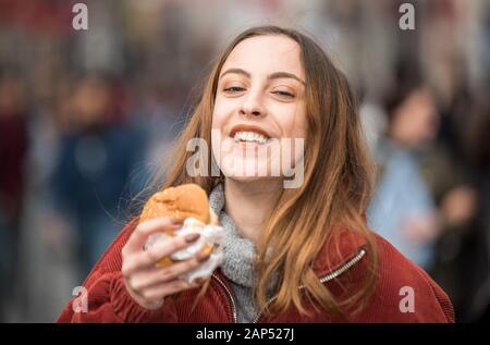 Beautiful young cute girl in fashionable clothes enjoys fast food hamburger whilewalking at a crowded street Stock Photo