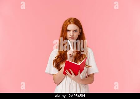 Serious-looking grumpy redhead woman frowning as thinking, pouting troubled, solving serious riddle, holding red notebook and touching chin with pen Stock Photo