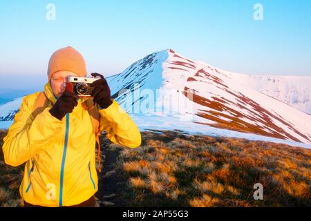 Photographer in yellow jacket taking photo with retro camera on snowy mountains Stock Photo