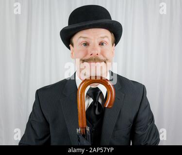 Portrait of Man in Dark Suit and Bowler Hat Smiling With Umbrella. Concept of Classic and Eccentric British Gentleman. Retro Fashion. Stock Photo