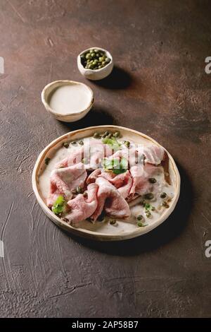 Vitello tonnato italian dish. Thin sliced veal with tuna sauce, capers and coriander served on ceramic plate over dark texture background. Stock Photo